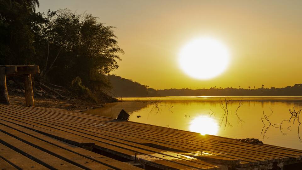 A wooden pier extending into a calm lake at sunset with trees on the left and a clear sky.