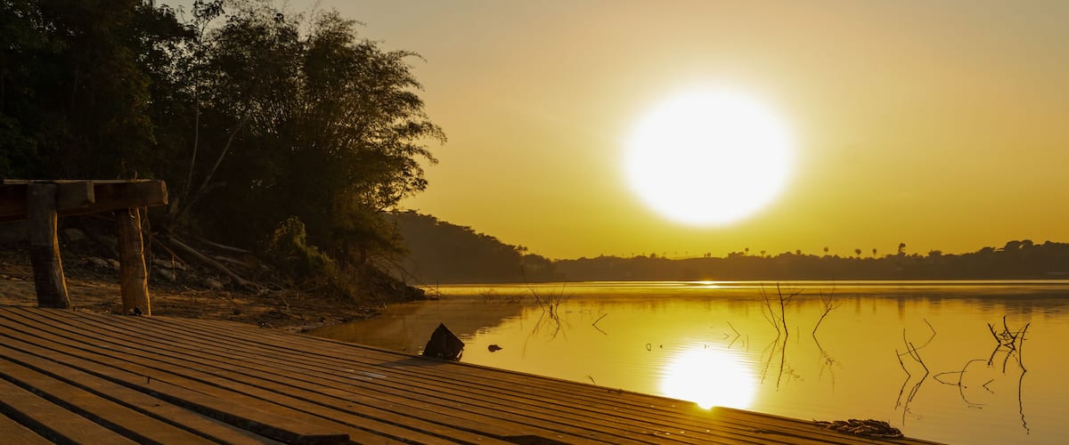 A wooden pier extending into a calm lake at sunset with trees on the left and a clear sky.