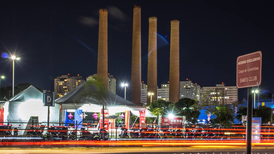 A cityscape at night with illuminated buildings, a large tent structure, and a sign reading "ACESSO EXCLUSIVO SAM'S CLUB."