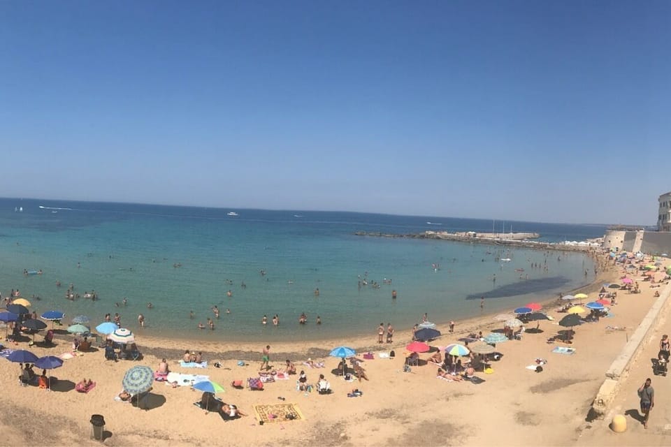 Uma praia cheia de gente, com pessoas se banhando e tomando sol, rodeada por edifícios e um céu azul claro.