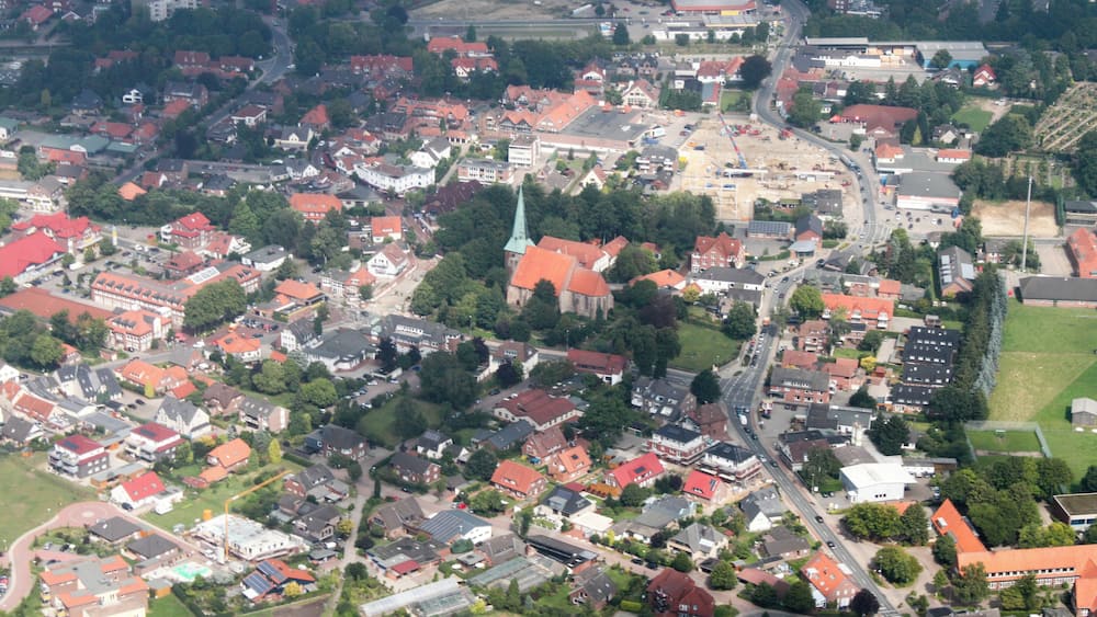 Une vue aérienne d’une ville avec des maisons aux toits rouges, une église dotée d’un clocher vert, et un mélange de bâtiments résidentiels et commerciaux.