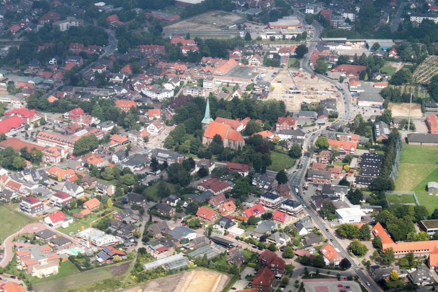 Vista aérea de un pueblo con casas de tejados rojos, una iglesia con campanario verde y una combinación de edificios residenciales y comerciales.
