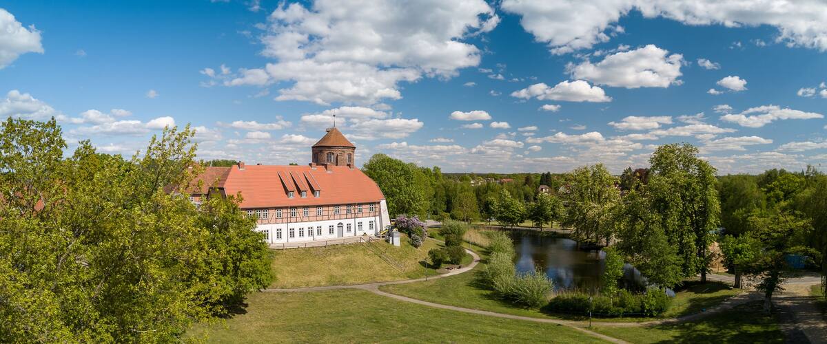 A large white building with a red roof and a tower, surrounded by trees and a lake.