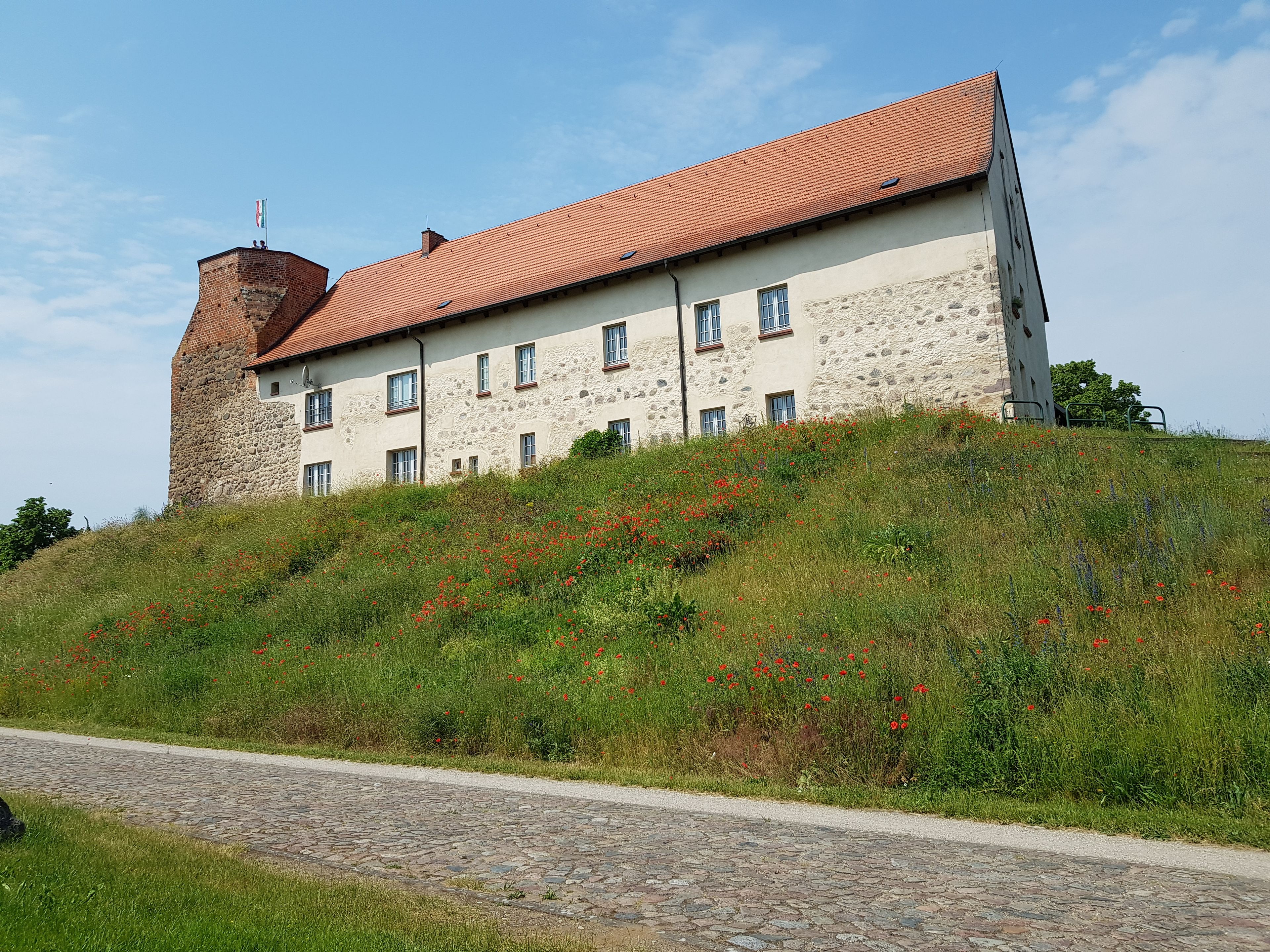 Ein historisches Gebäude mit rotem Ziegeldach und Steinmauern, gelegen auf einem grasbewachsen Hügel mit einem Pflasterweg, der hinaufführt.