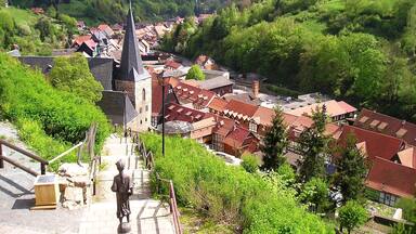 A town with a prominent church, red-roofed buildings, and lush green hills in the background.