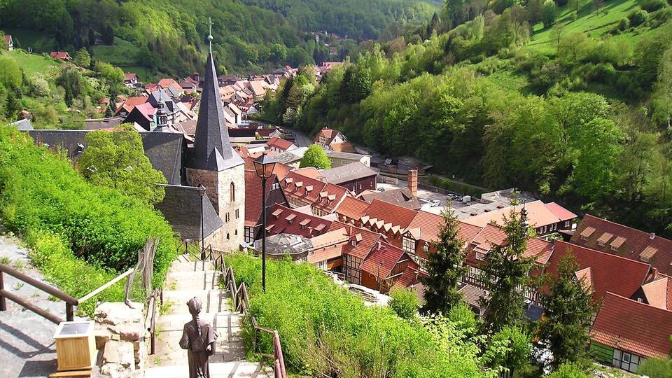 A town with a prominent church, red-roofed buildings, and lush green hills in the background.