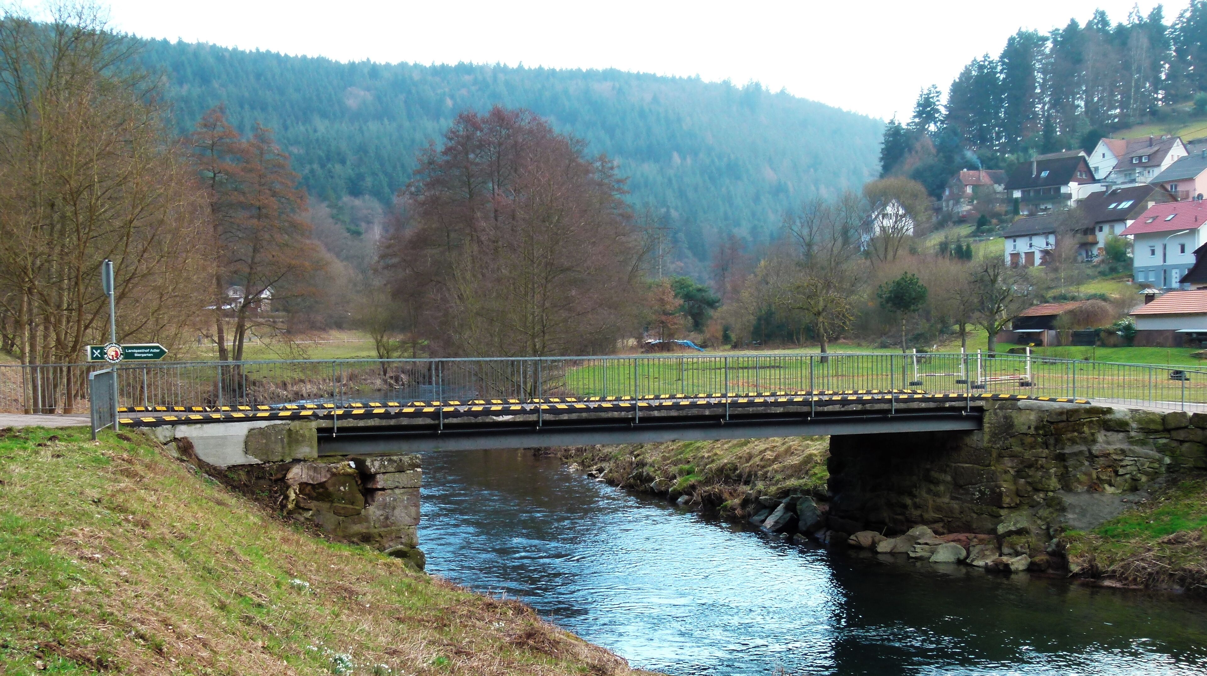 A stone bridge over a river with a metal railing, surrounded by trees and houses in the background.
