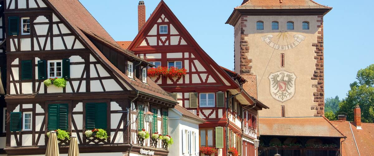 A traditional half-timbered house with green shutters and flower boxes, a church tower with a clock, and a clear blue sky.