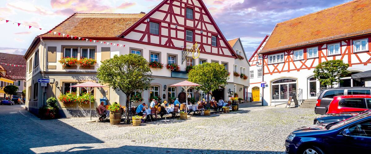 A traditional European town square with half-timbered buildings, outdoor seating, and cobblestone streets.