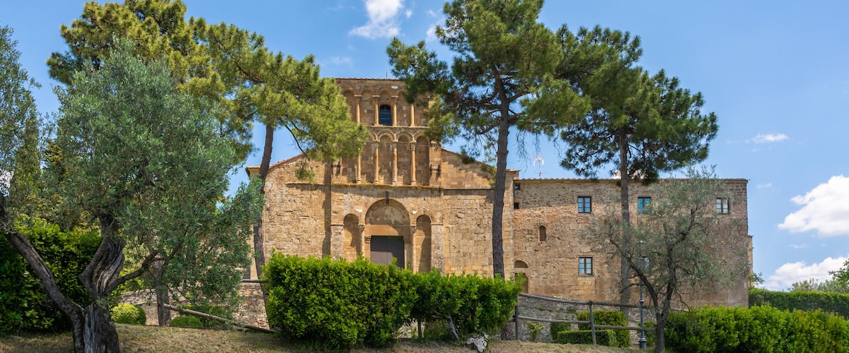 A historic stone building with arched windows and a bell tower, surrounded by trees and shrubs.