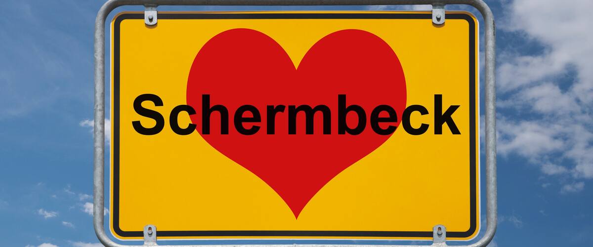 A yellow sign with a red heart and the word "Schermbeck" against a blue sky with clouds.