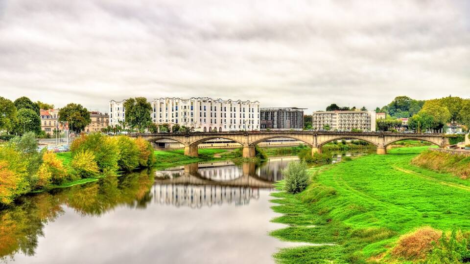 A river with a bridge, green fields, and buildings in the background.