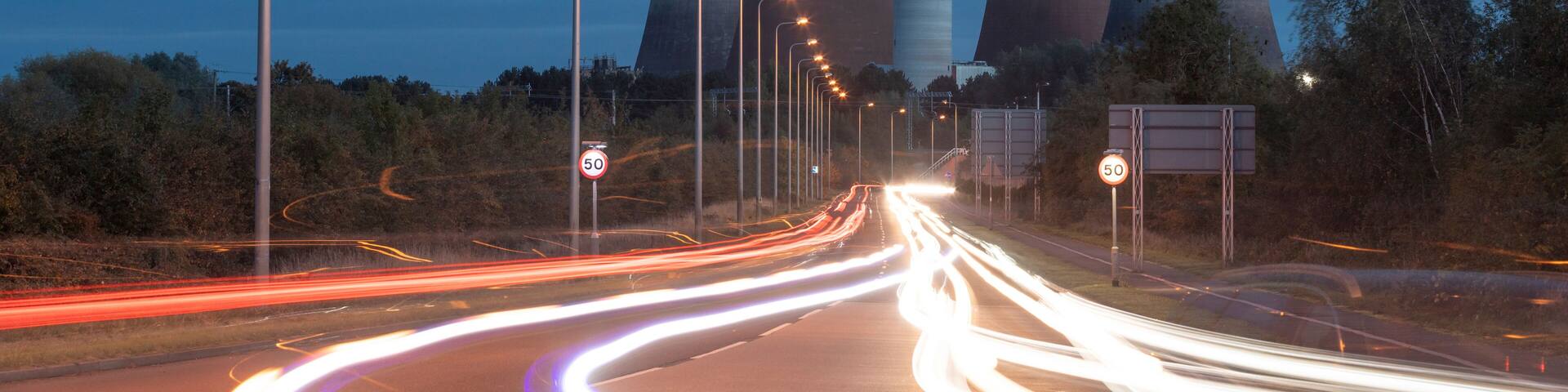 UK, England, Rugeley, Vehicle light trails stretching along illuminated road at dusk with cooling towers in background