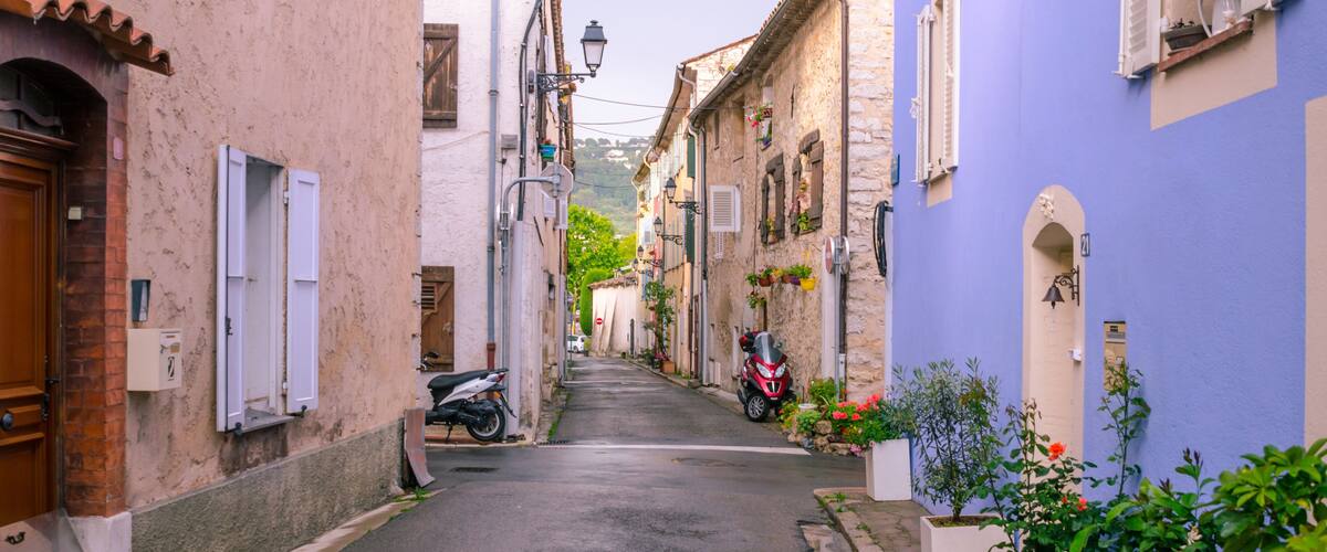 A narrow street with buildings painted in pastel colors, a parked motorcycle, and potted plants along the sides.