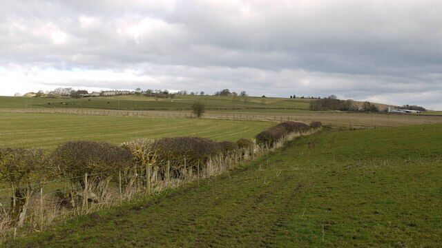 A rural landscape with open fields, a fence, and distant trees under a cloudy sky.