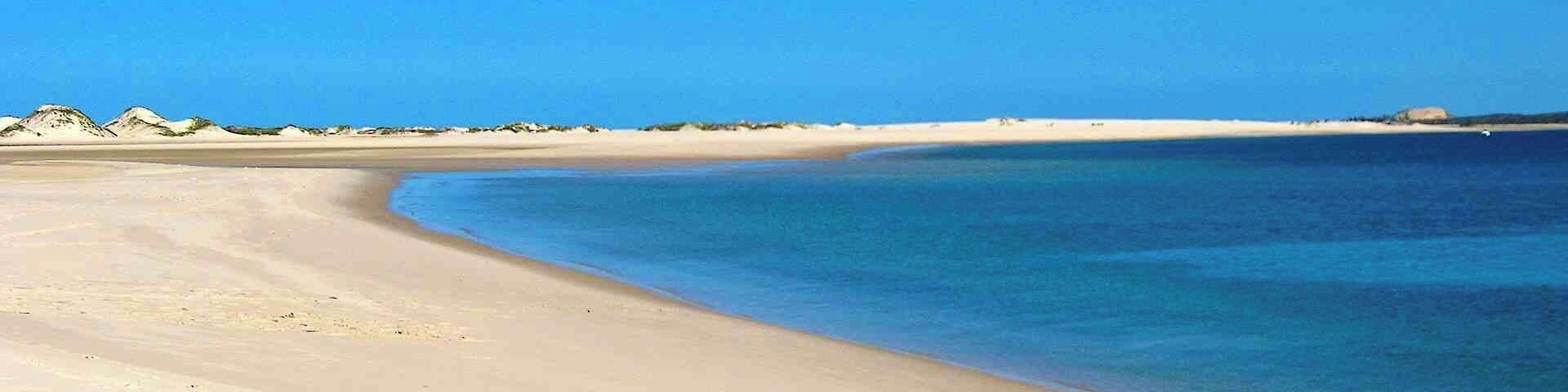 A sandy beach with clear blue water and distant dunes.
