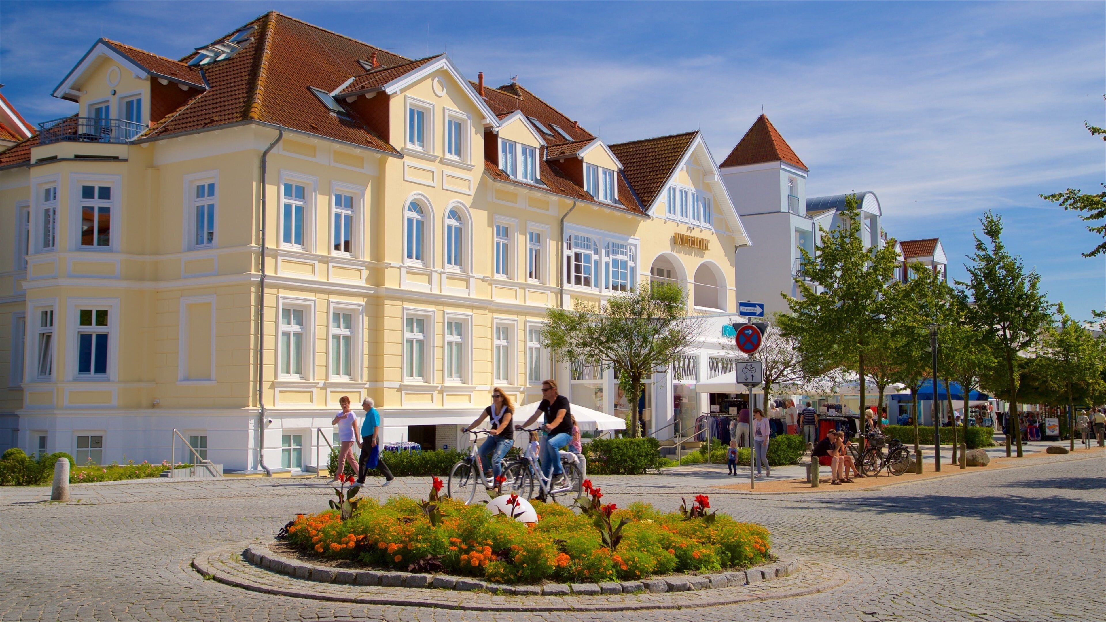 A cobblestone square with a yellow building, people on bicycles, and a flower garden.