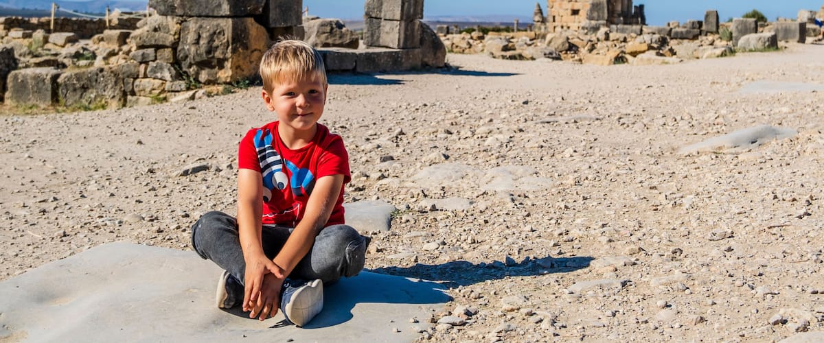 A boy sitting on a rocky surface with ancient ruins and a clear sky in the background.