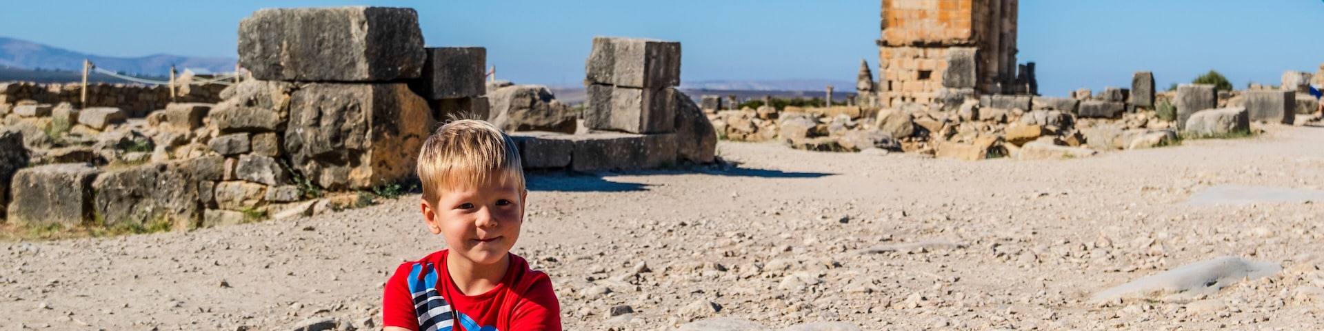 A boy sitting on a rocky surface with ancient ruins and a clear sky in the background.