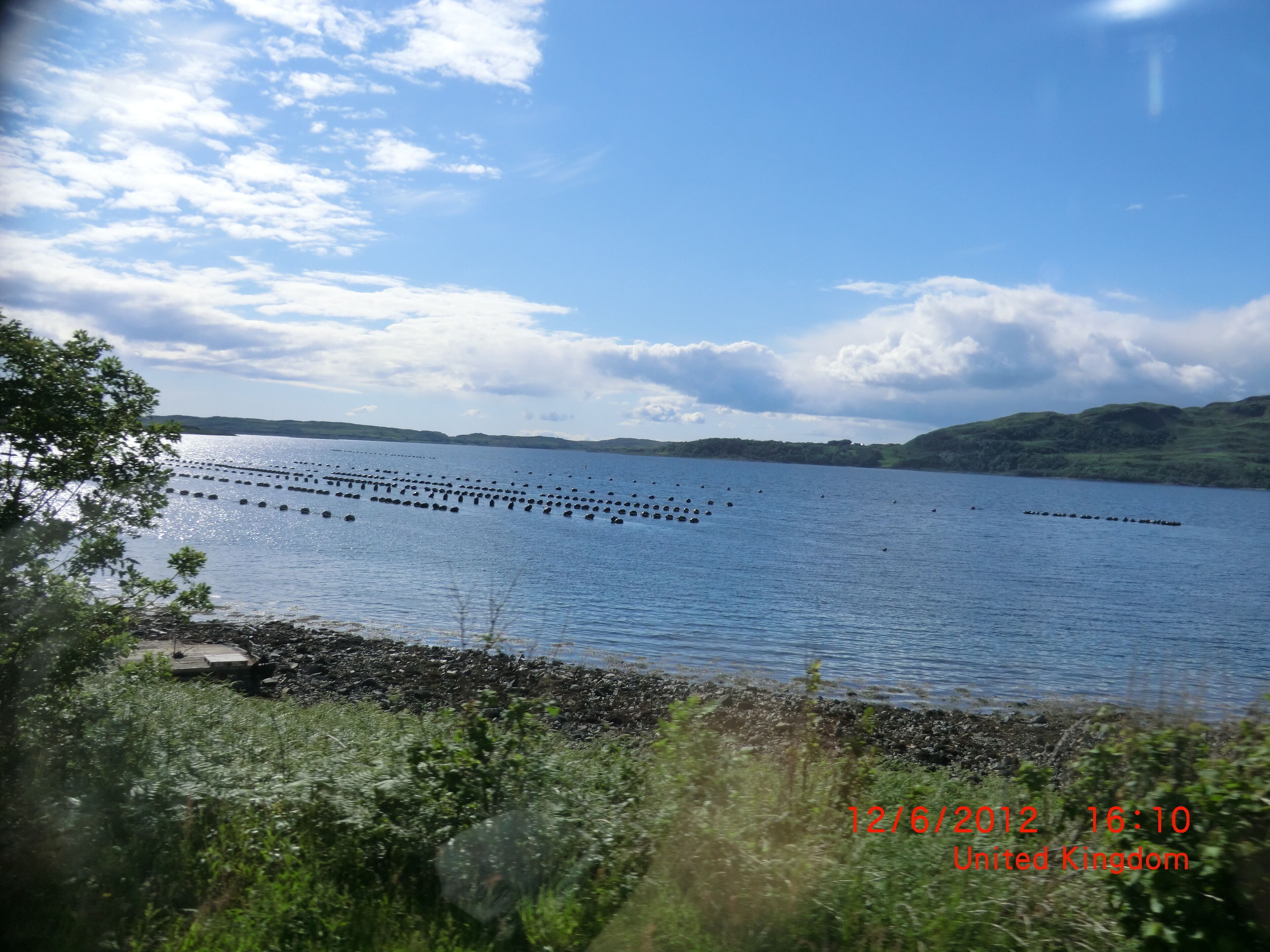 A calm lake with floating buoys, surrounded by hills and greenery.