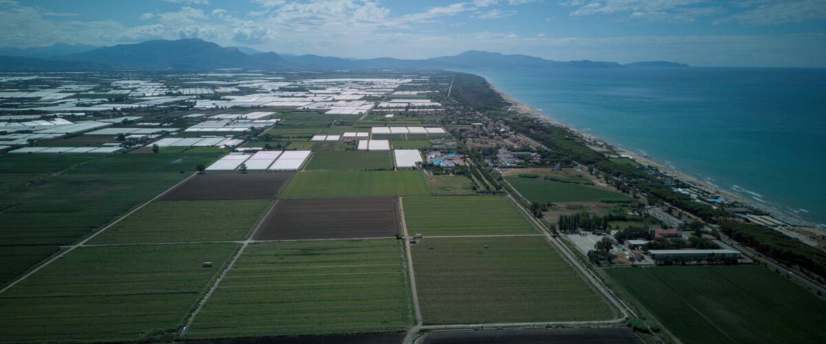 An aerial view of a coastal agricultural area with fields, a beach, and mountains in the background.