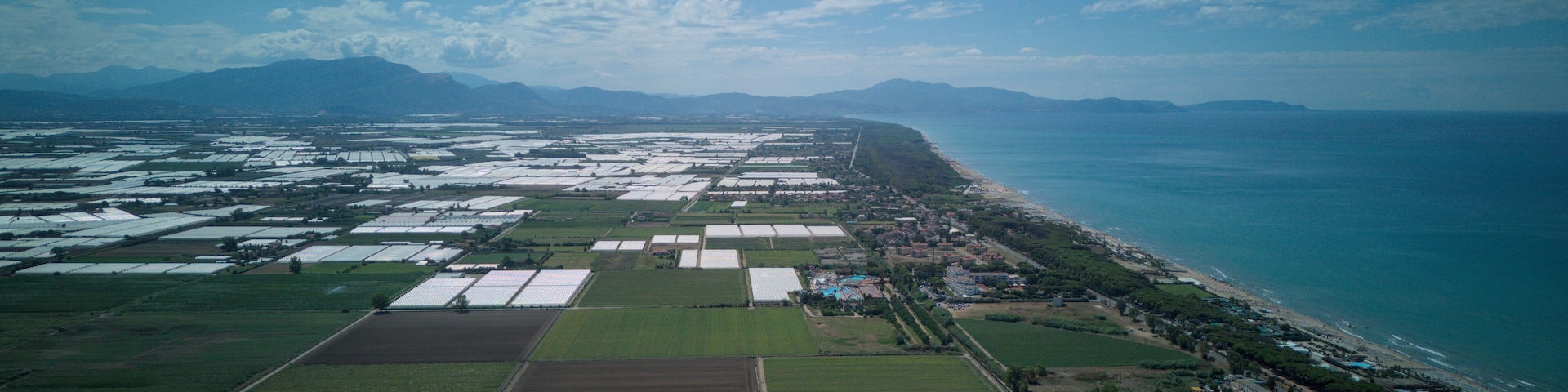 An aerial view of a coastal agricultural area with fields, a beach, and mountains in the background.