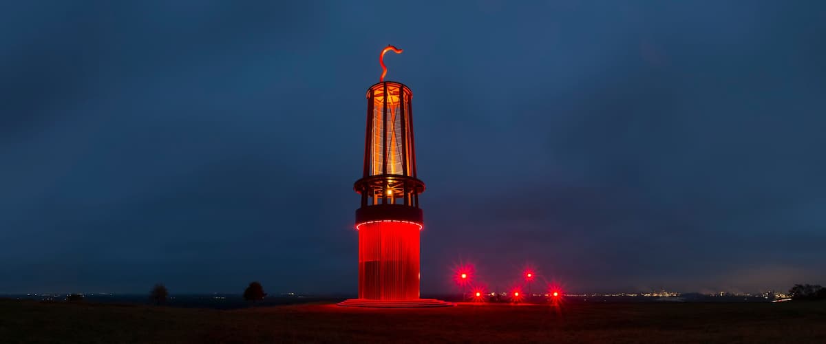 A tall tower with a flame at the top, surrounded by a field with distant city lights.