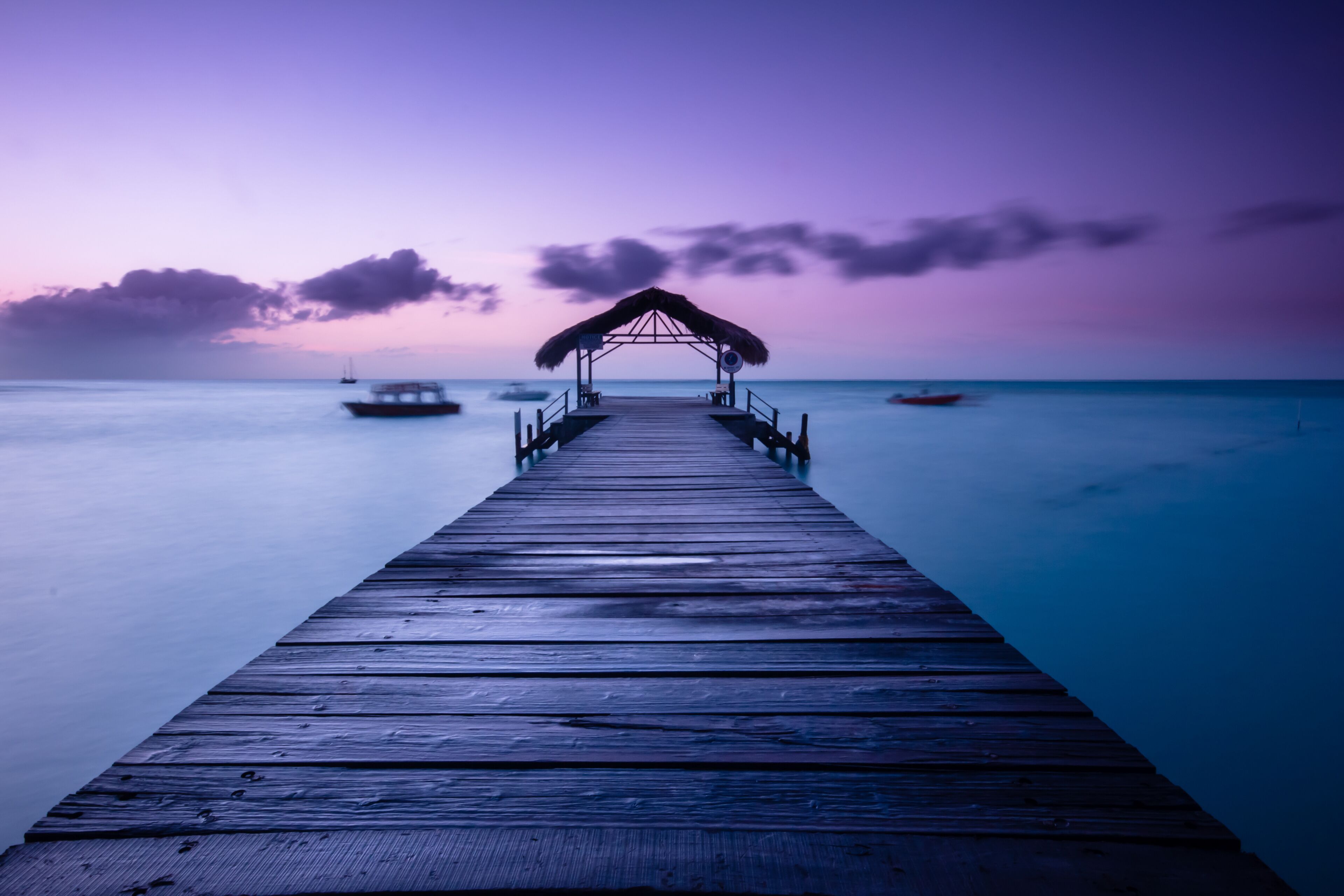 A wooden pier extending into calm waters, with a thatched-roof structure at the end and boats in the distance.