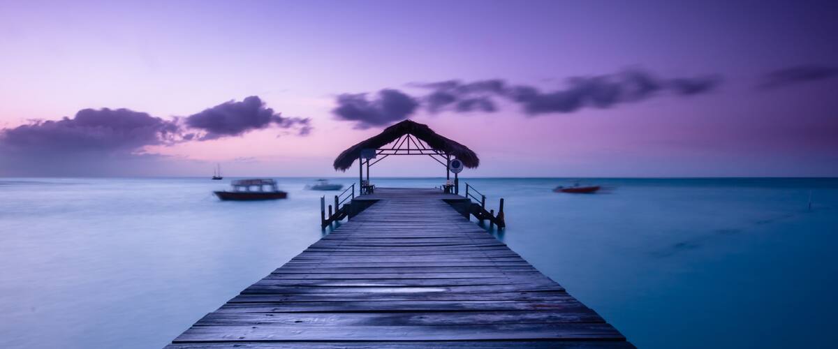 A wooden pier extending into calm waters, with a thatched-roof structure at the end and boats in the distance.