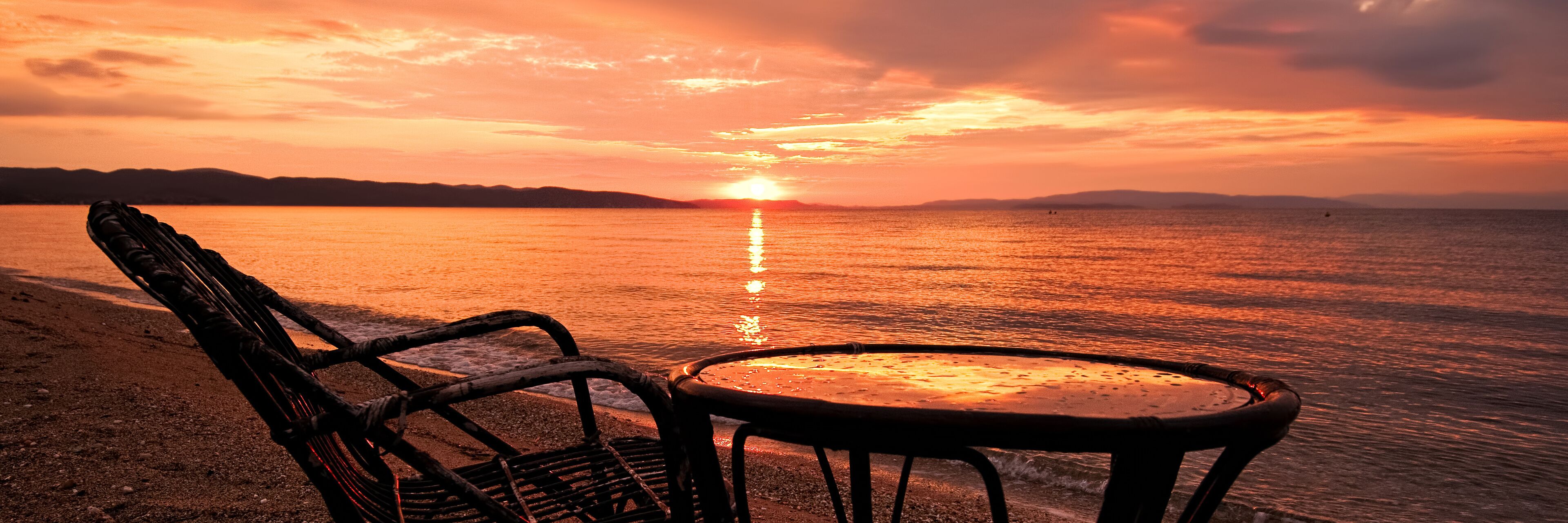 A beach chair and table facing a sunset over water.