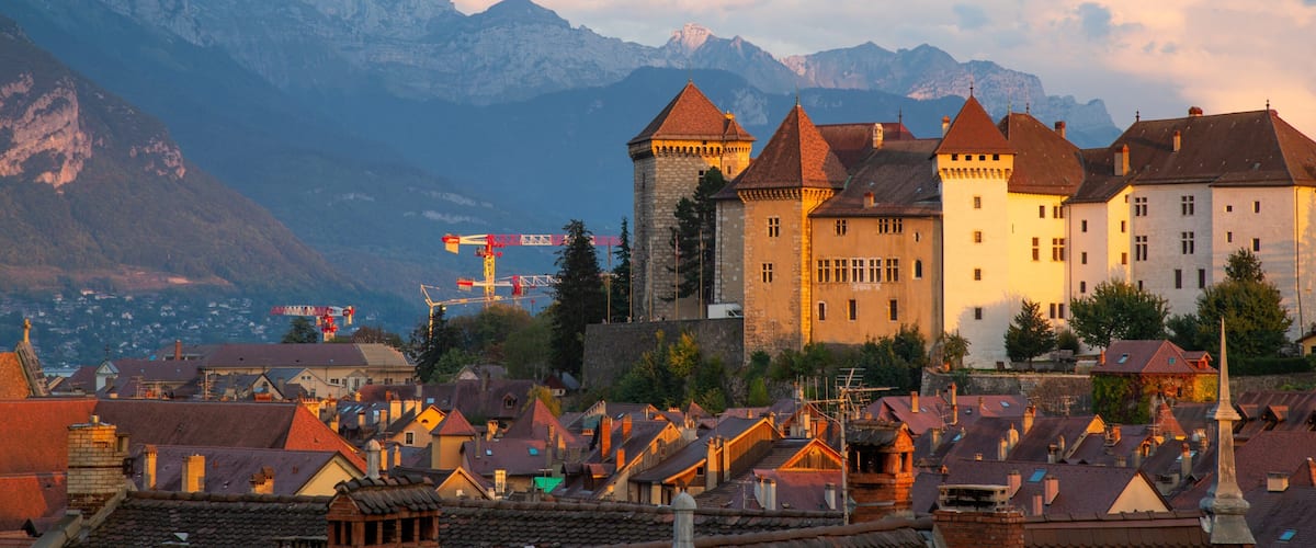 A historic town with medieval architecture, including a prominent tower and castle, set against a backdrop of mountains and a bridge.