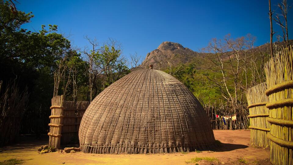 A traditional thatched hut surrounded by bamboo fences and trees, with a mountain in the background.