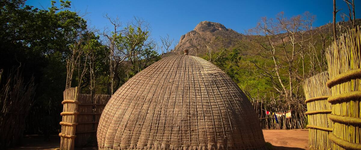 A traditional thatched hut surrounded by bamboo fences and trees, with a mountain in the background.