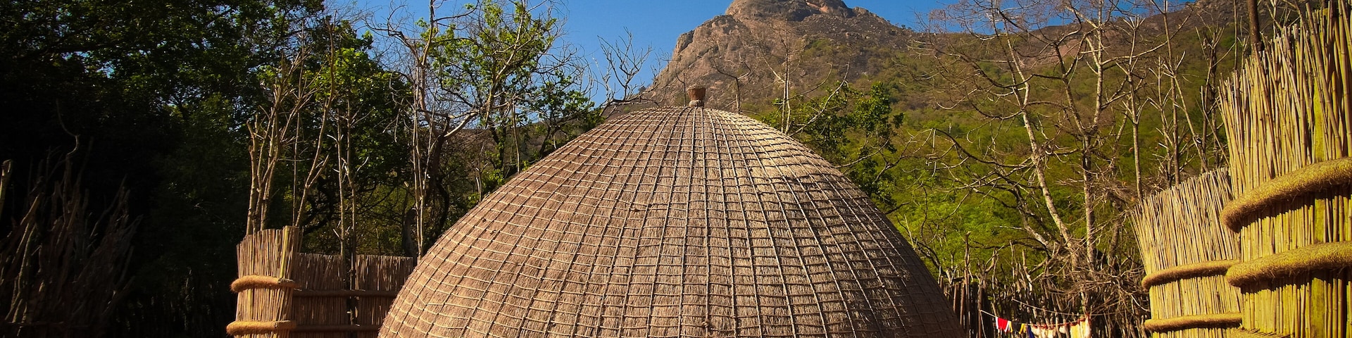 A traditional thatched hut surrounded by bamboo fences and trees, with a mountain in the background.