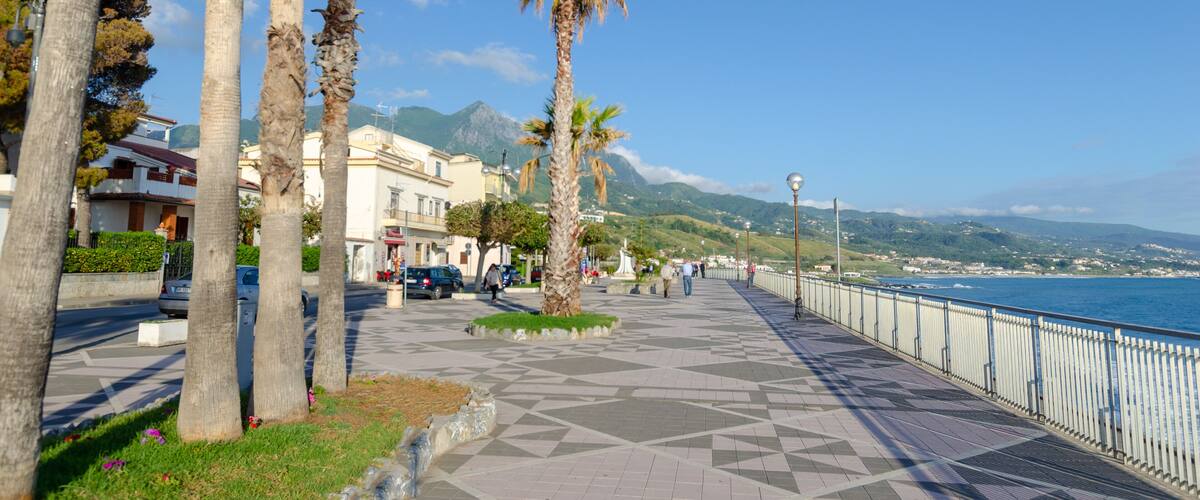 A coastal promenade with palm trees, a tiled walkway, and a view of the sea and mountains.