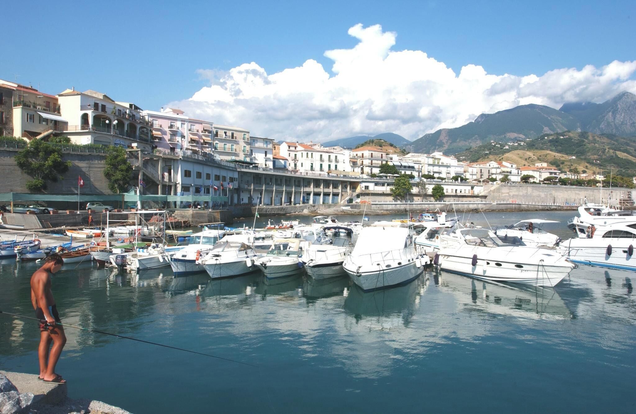 A marina with boats docked, a person fishing, and buildings along the waterfront.