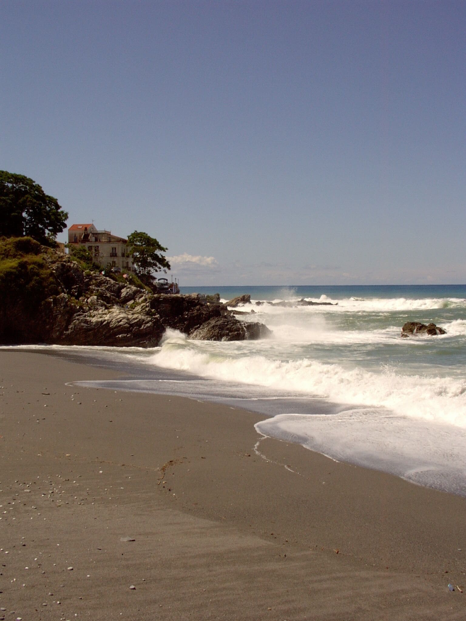 A beach with waves crashing onto the shore, a rocky outcrop with a building, and a clear blue sky.