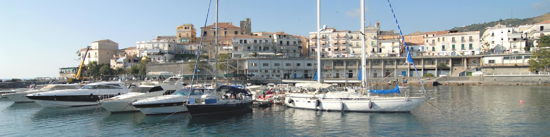 A marina with several sailboats docked, a clear blue sky, and buildings in the background.