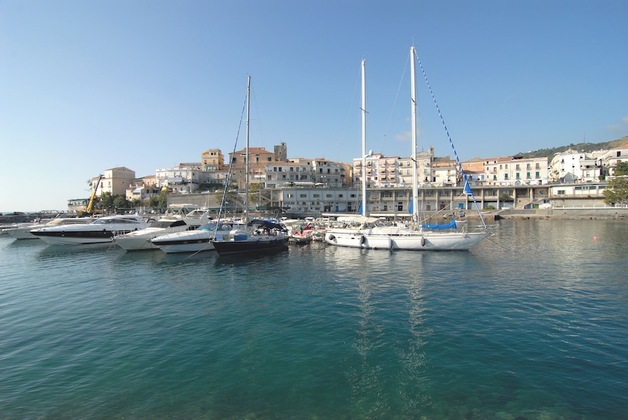 A marina with several sailboats docked, a clear blue sky, and buildings in the background.