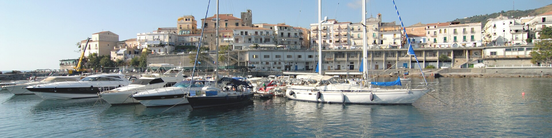 Un puerto deportivo con varios veleros amarrados, cielo azul despejado y edificios al fondo.