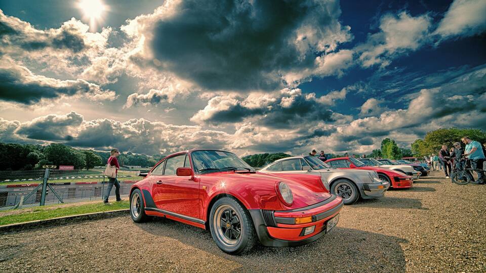 A red sports car parked on a gravel lot with a cloudy sky in the background.