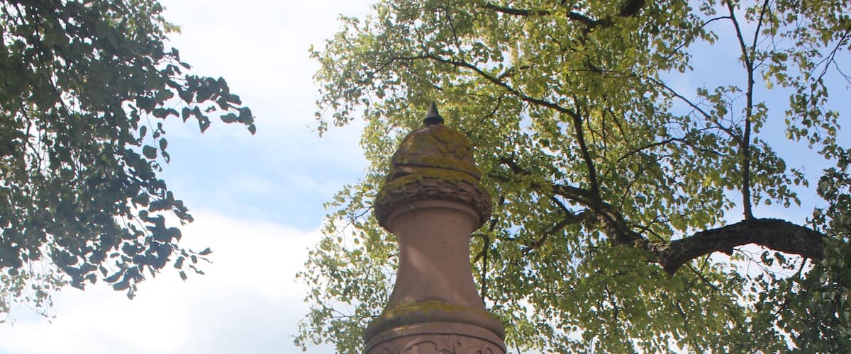 A tall, pink monument with inscriptions in a foreign language, surrounded by a field and trees.