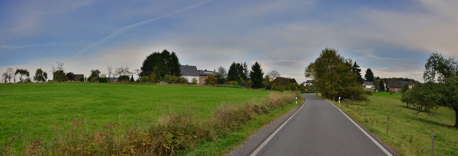 Eine ländliche Straße, gesäumt von Feldern und Bäumen, führt zu Häusern in der Ferne.