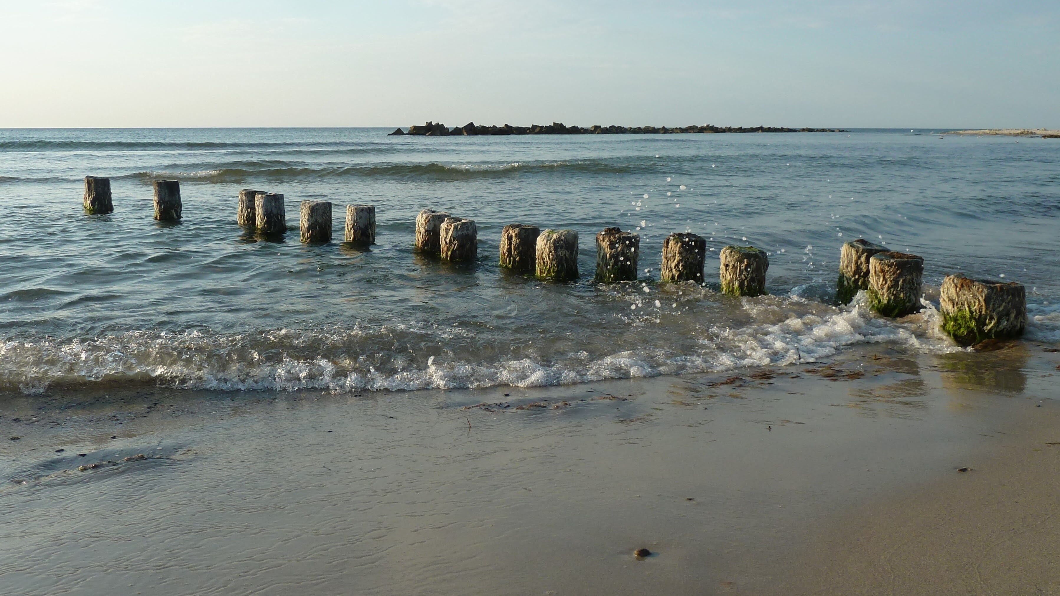 A sandy beach with wooden posts in the water and a distant rocky outcrop.