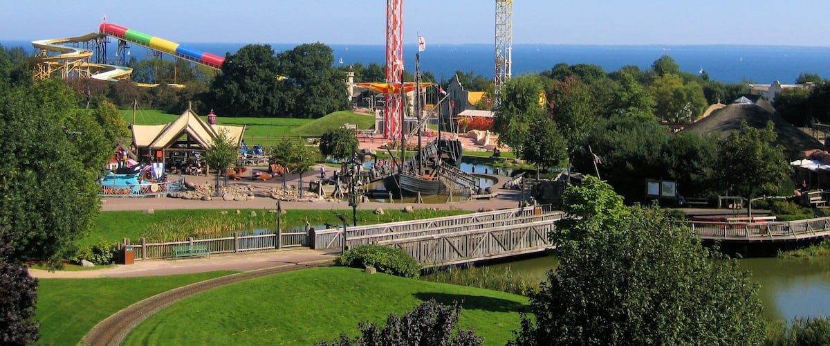Un parc temàtic amb una alta muntanya russa, un tobogan d'aigua i un pont sobre un estany.