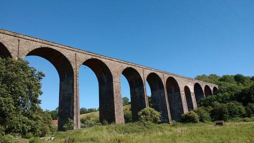 Eine lange Viaduktbogenbrücke überspannt ein Tal, umgeben von üppiger Vegetation und blauem Himmel.