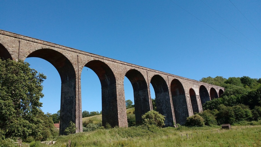 Un largo viaducto con múltiples arcos que cruza un valle, rodeado de abundante vegetación y un cielo azul despejado.