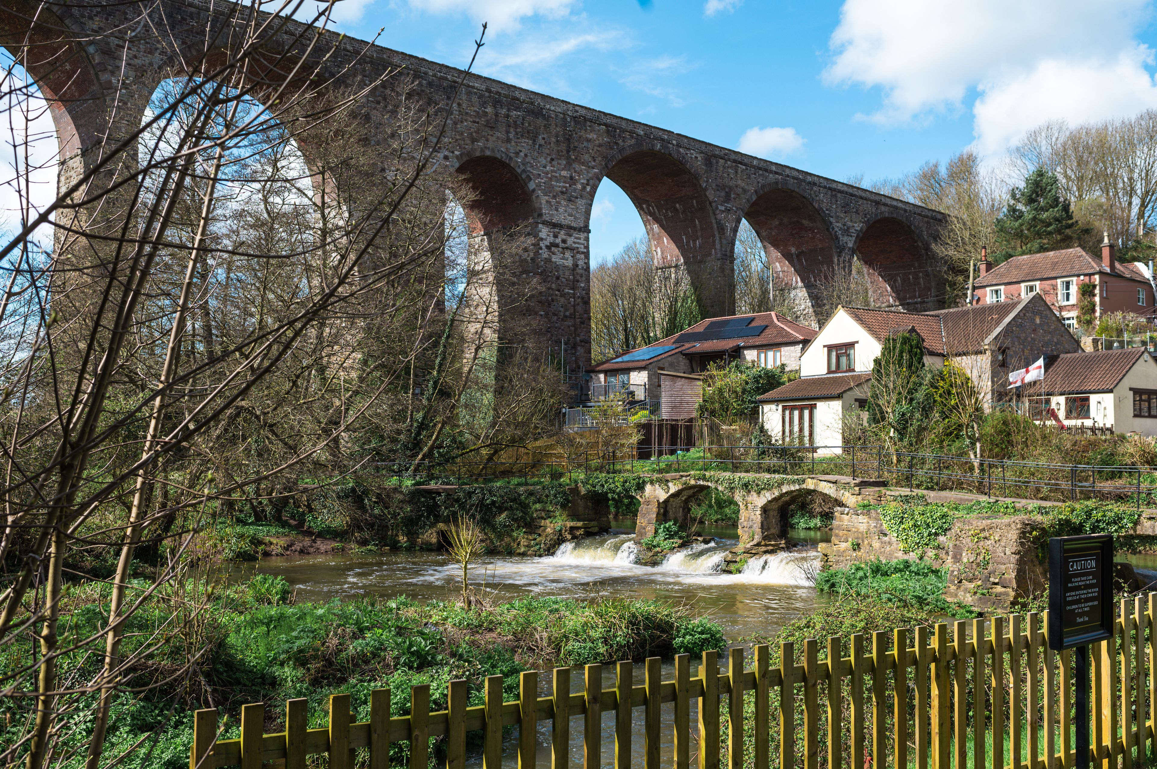 Eine Steinbrücke mit mehreren Bögen über einem Fluss, umgeben von Häusern und Grünflächen.