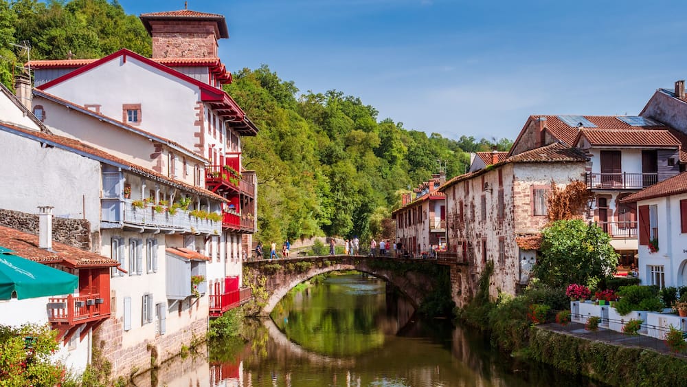 Une rivière traversée par un pont de pierre, des bâtiments dotés de balcons et, au loin, une végétation luxuriante.