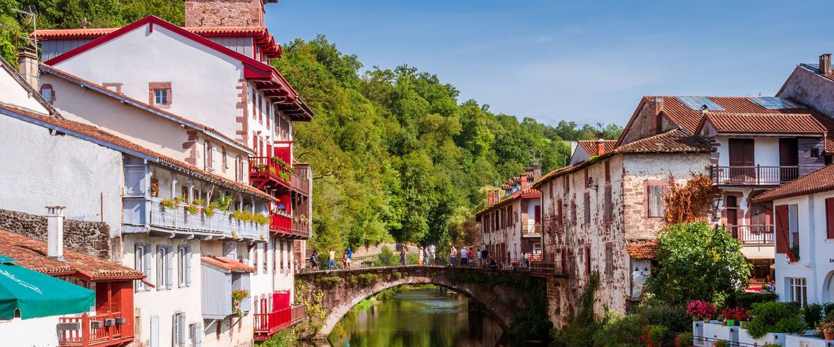 A river with a stone bridge, buildings with balconies, and lush greenery in the background.